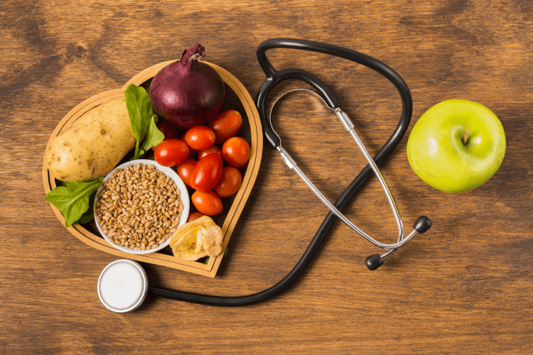 Image présentant un bol en forme de coeur, remplie de fruits, de légumes et de graines, située à gauche. A côté de ce bol, nous pouvons visualiser un stéthoscope ainsi qu'une pomme positionnée à droite du stéthoscope.